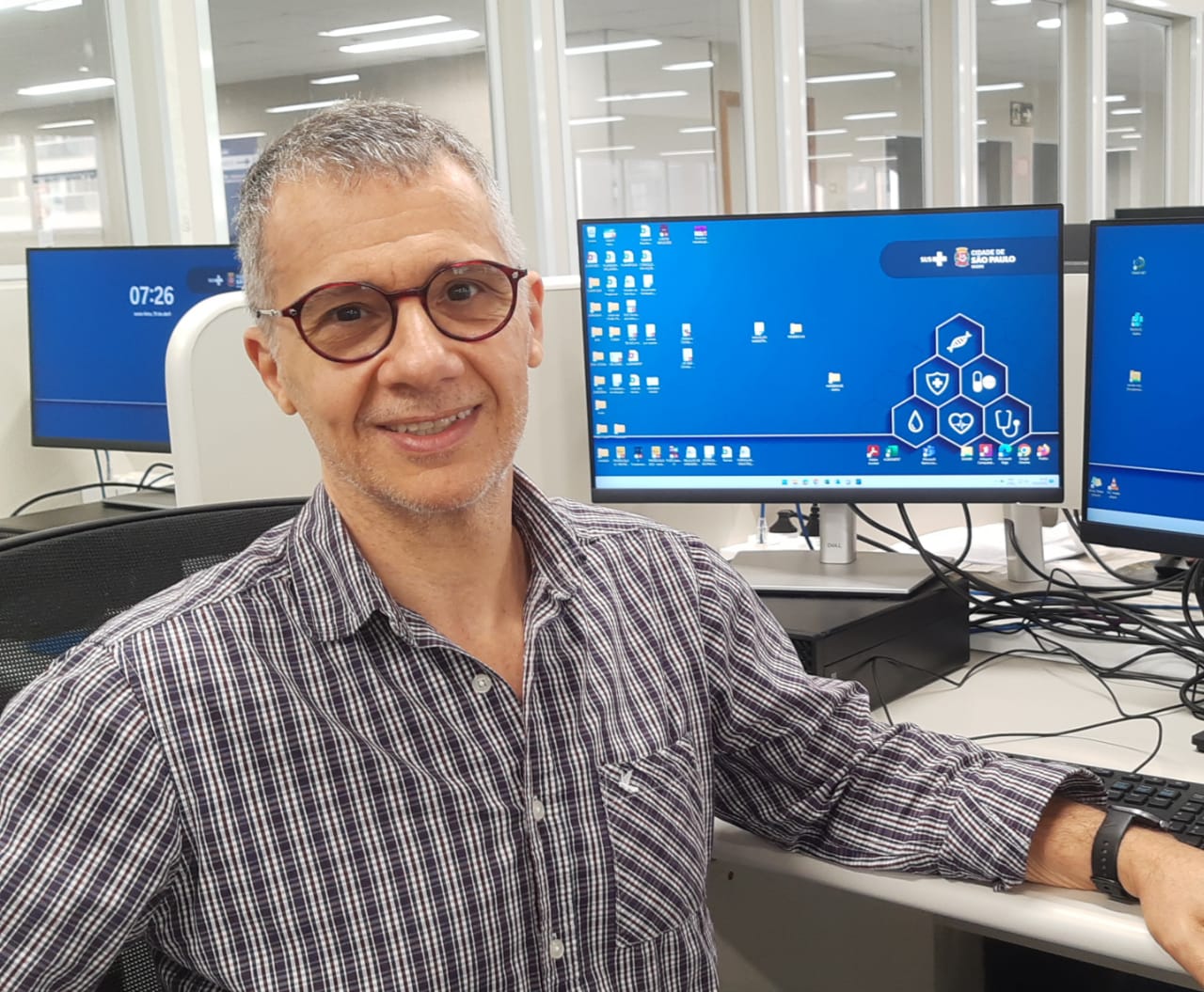 A foto mostra Carlos, um homem branco de cabelos grisalhos e óculos, em frente a um computador, no seu locald e trabalho. 